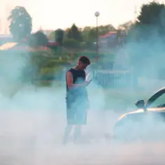 Młody mężczyzna z telefonem w dłoni stoi w kłębach dymu. Obok niego samochód, z którego **diesel dymi na czarno**.