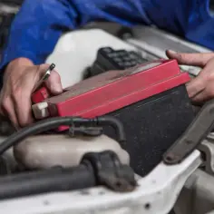 Mechanik wymienia akumulator renault megane 3. Czerwona pokrywa baterii jest widoczna, a ręce trzymają narzędzie.