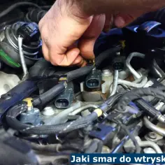 Dłoń mechanika pracuje przy silniku, podłączając wtryskiwacze. Kluczowe jest użycie odpowiedniego smaru do wtryskiwaczy, by zapewnić ich prawidłowe działanie.