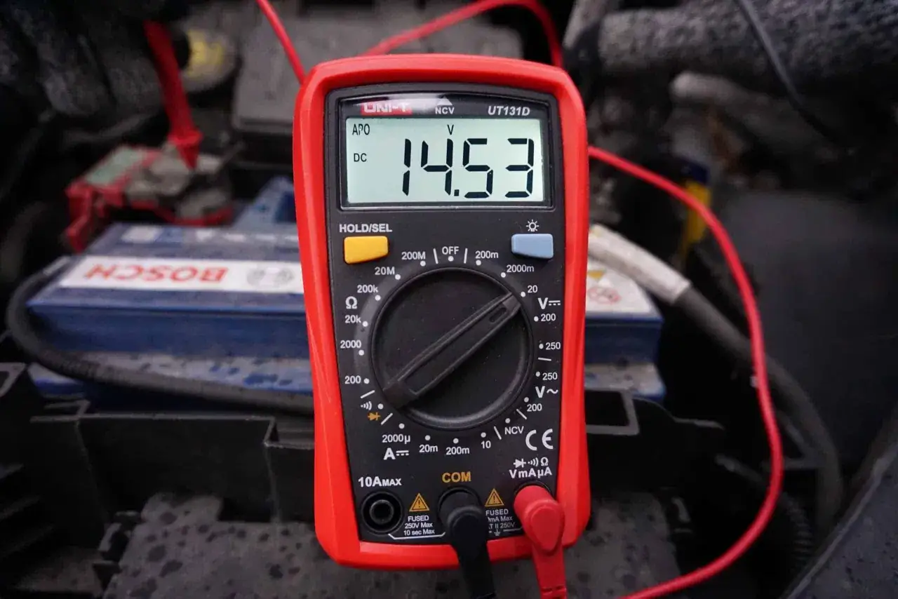 Multimetr pokazuje 14.53V, co jest kluczowe, jak sprawdzić alternator miernikiem. W tle widać akumulator samochodowy.